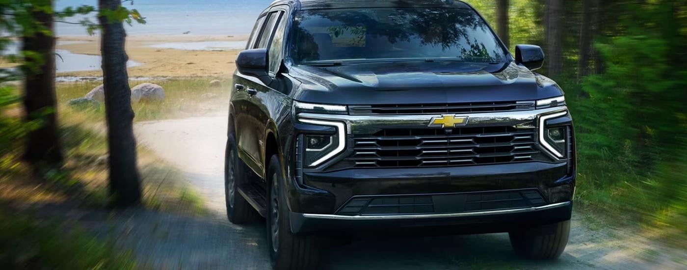 A black 2025 Chevy Tahoe is driving on a gravel road lined with trees.